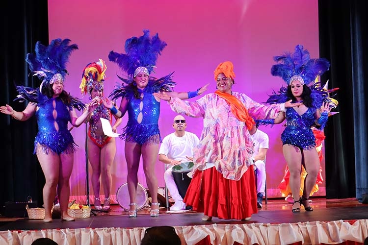 Performers in blue sequined costumes and a woman in a colorful dress on stage.