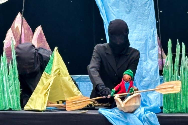 Two black-clad puppeteers perform a puppet show with a small boat and a yellow tent.