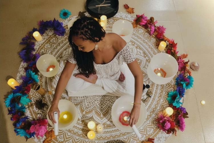 A woman in white sitting cross-legged on a patterned rug, surrounded by candles and singing bowls.