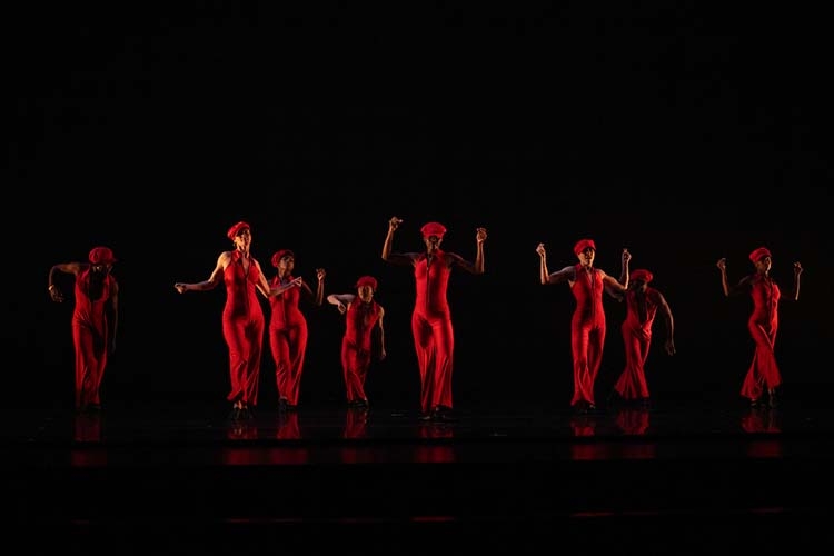 Group of dancers in red outfits and hats performing on a dark stage.