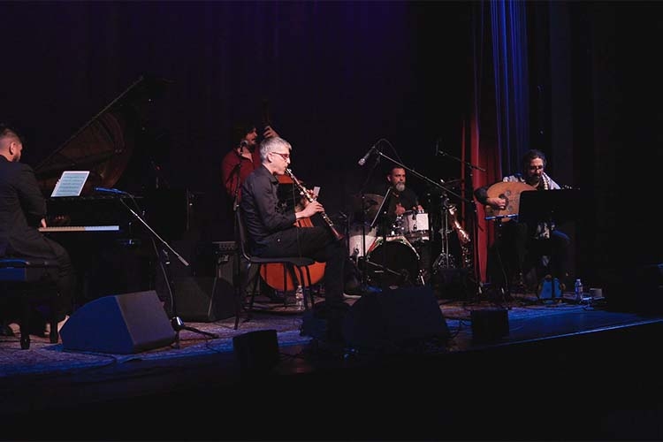 Photograph of a jazz band performing on a dark stage with instruments including a piano and oud.