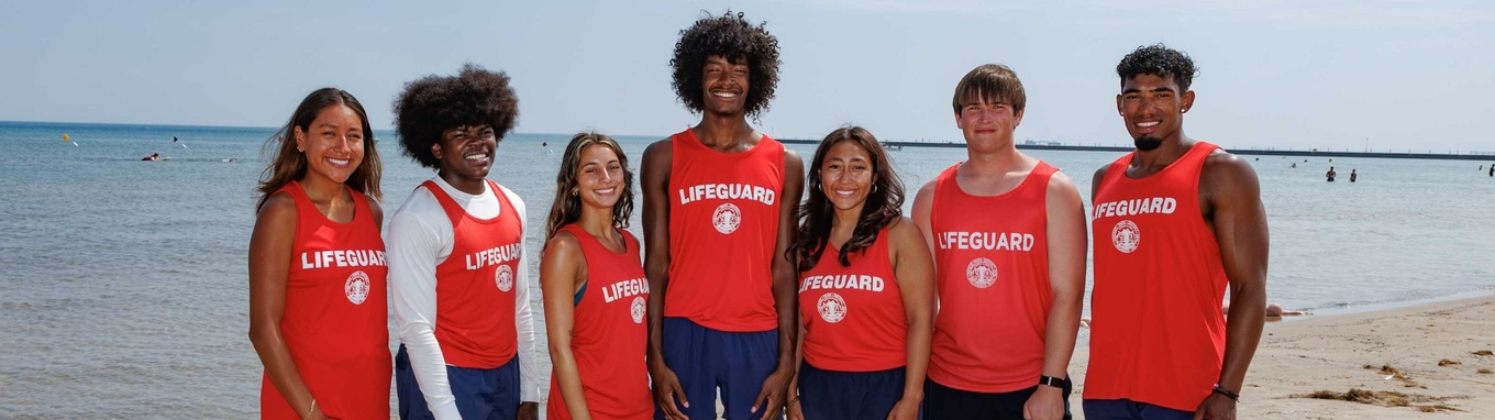 Photograph of a diverse group of seven lifeguards in red tank tops standing on a beach.