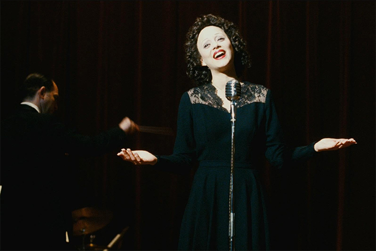 A photograph of a woman with curly hair singing into a vintage microphone on stage.