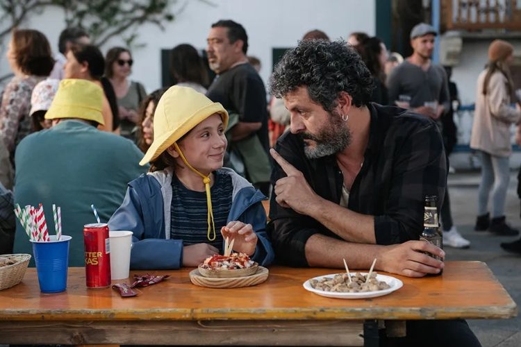 A man with a beard gestures while talking to a young child in a yellow hat at an outdoor table.