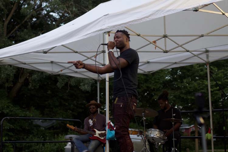 A Black male vocalist performs on an outdoor stage with a guitarist and drummer.