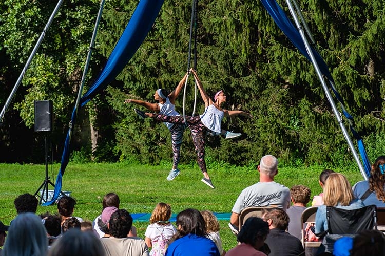 Two aerialists perform on a hoop outdoors in front of a seated audience.
