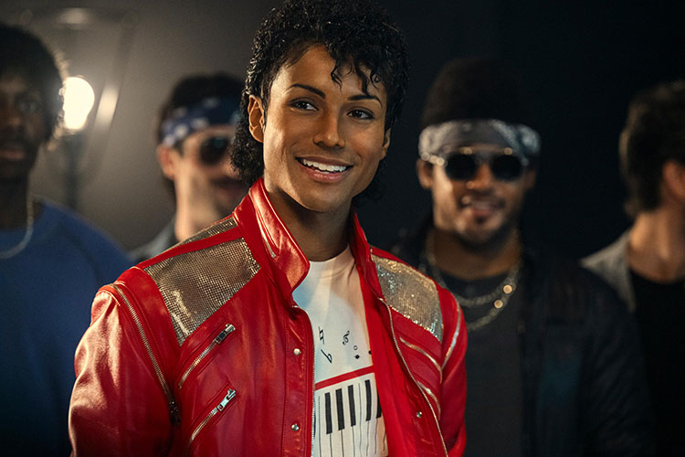 A smiling man dressed as Michael Jackson in a red jacket surrounded by background dancers.