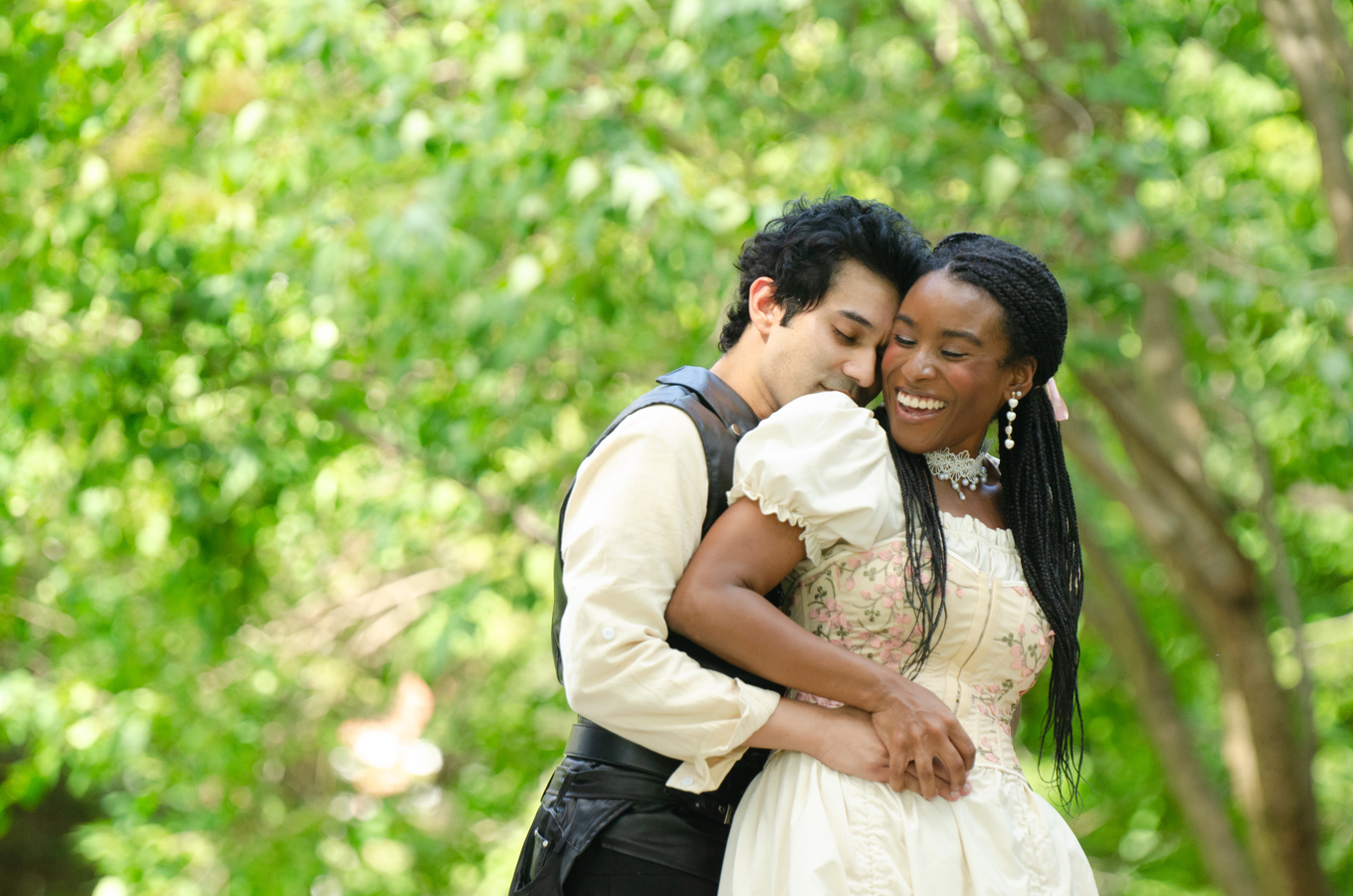 A man in a rustic vest and a woman in a floral Victorian-style dress embrace in a lush green park.