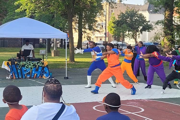 Performers dressed as Dragon Ball Z characters dancing on a basketball court outdoors.