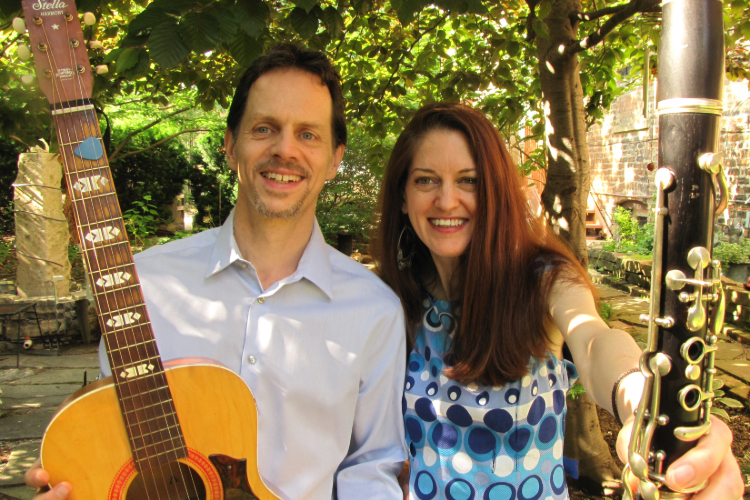 A man holding an acoustic guitar and a woman holding a clarinet smile in a garden.