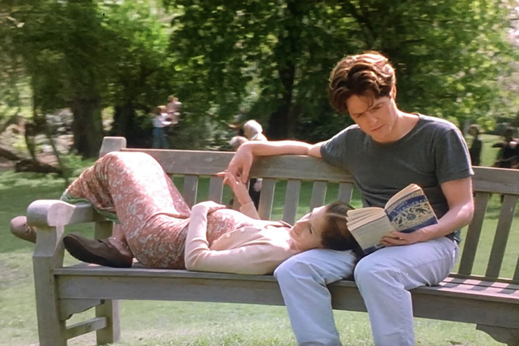A man reads a book while a woman lies with her head in his lap on a park bench.
