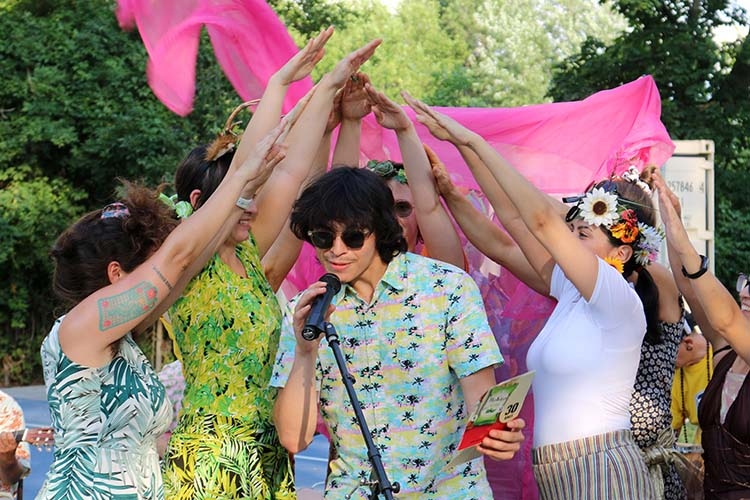 A group of people wearing floral attire and flower crowns surround a singer, forming an arch.