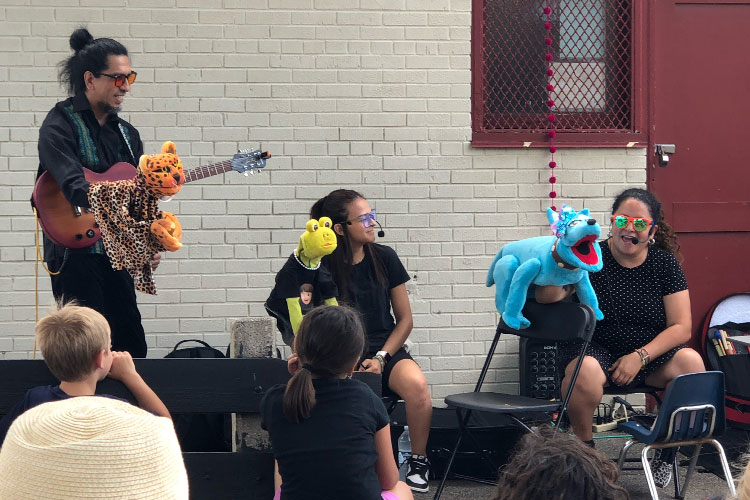 A guitarist and two puppeteers perform for children outdoors.