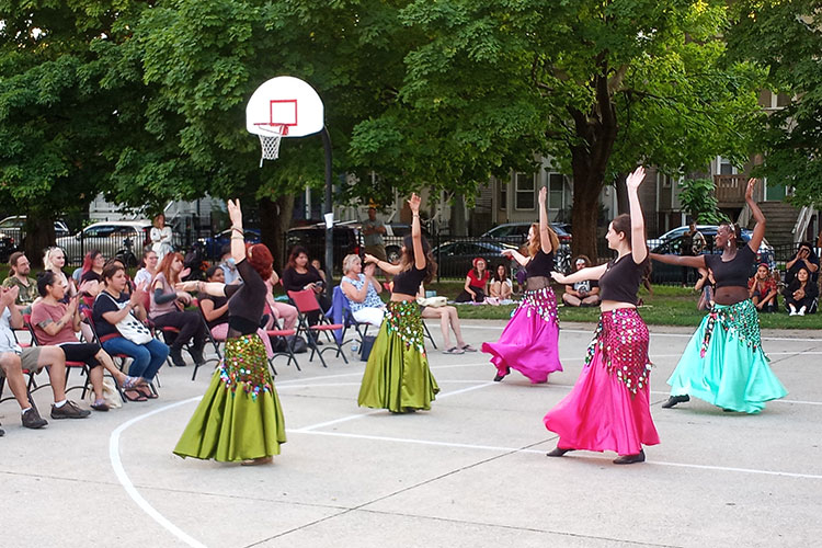 Four dancers in long skirts and black tops perform on a basketball court for an audience.