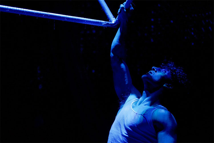 Photograph of a performer in a blue-lit stage performance hanging from a bar.