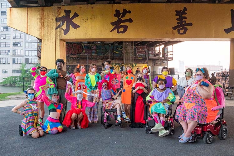 A diverse group of performers in colorful costumes posing outdoors under a concrete bridge.