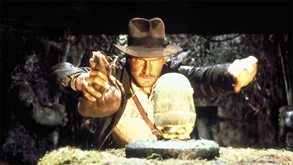 Indiana Jones in a fedora carefully replaces a golden idol with a sandbag in a dark cave.