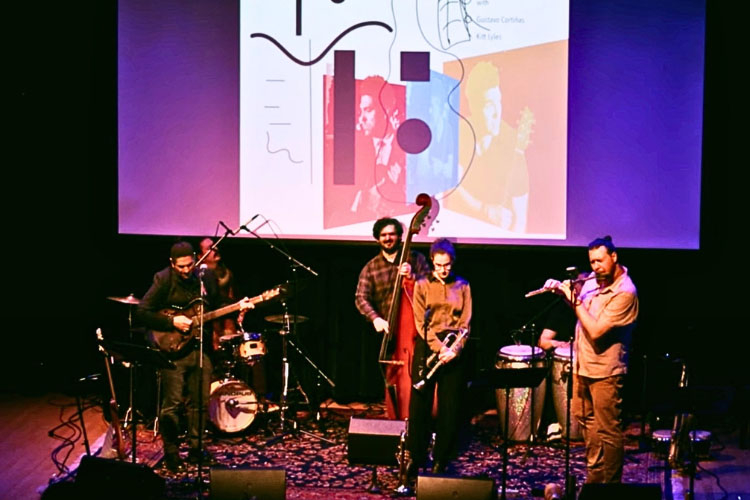 A jazz quartet performs on stage in front of a promotional screen.