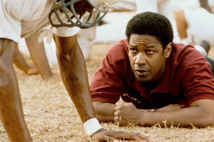 Denzel Washington as a coach lying on the grass next to a football player in a film still.