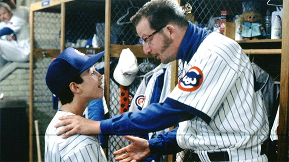 A baseball coach in a Cubs uniform talks to a young player in a locker room.