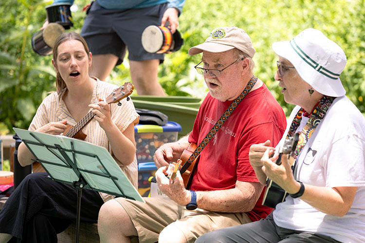 Three people playing ukuleles and singing outdoors in a garden setting.