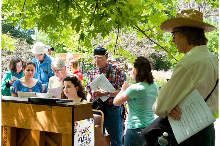 A group of people practicing music outdoors around a wooden piano under a large leafy tree.
