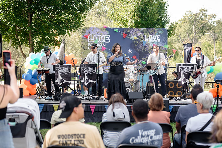 A band performs on an outdoor stage at a park, viewed from the audience.