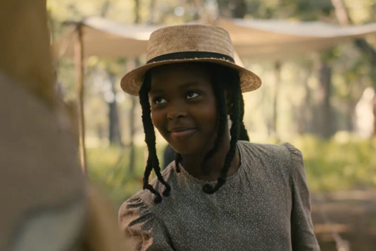 Young girl wearing a straw hat and floral-print dress, smiling in a wooded area.