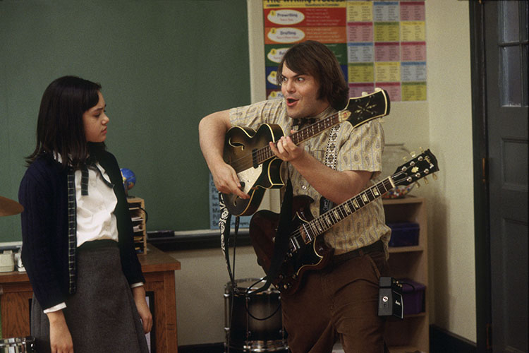 Jack Black plays two electric guitars in a classroom while a student looks on.