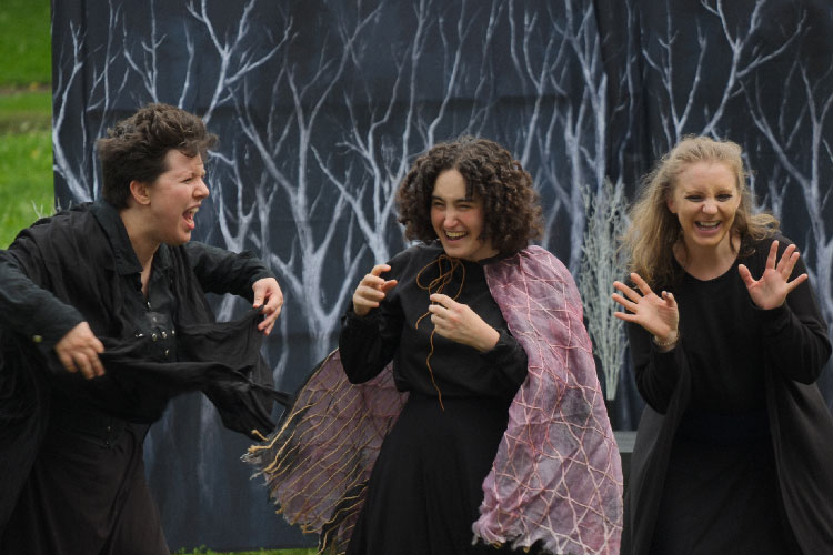 Three actors in black costumes perform on stage in front of a dark, wintry forest backdrop.