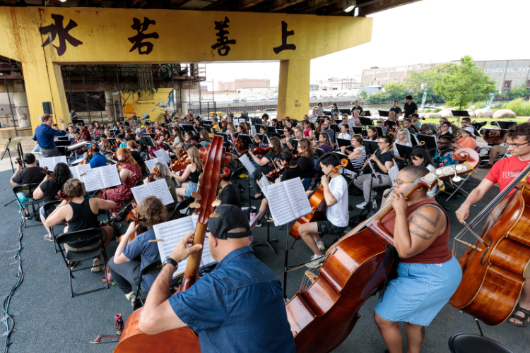 Photograph of a large, diverse community orchestra performing under a concrete bridge.