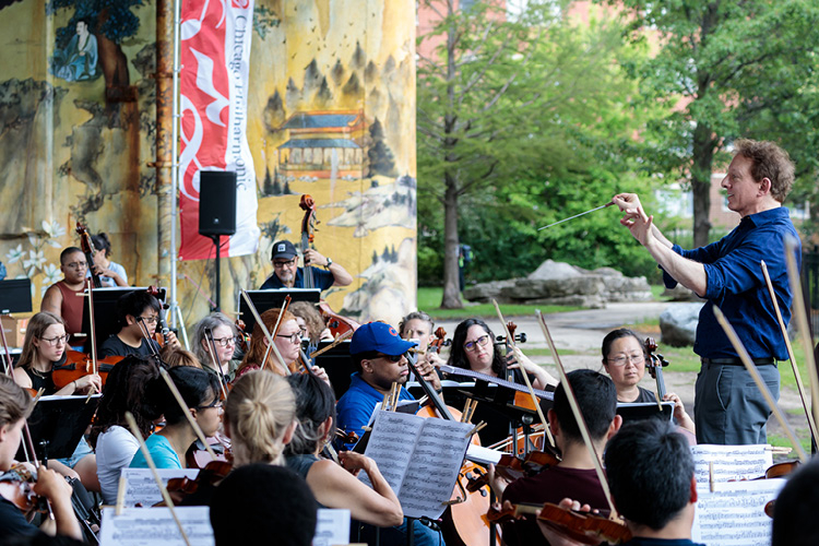 A male conductor leads an outdoor orchestra in front of a painted mural.