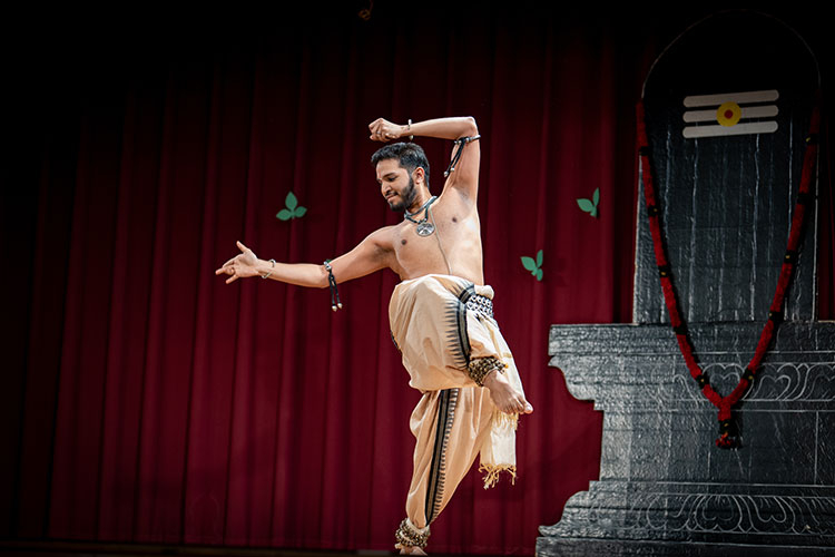 A male dancer in traditional attire performs on stage in front of a red curtain and stone sculpture.