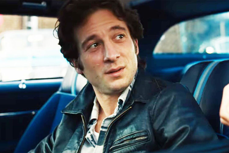 A young man with dark, wavy hair sits in a car wearing a leather jacket over a plaid shirt.