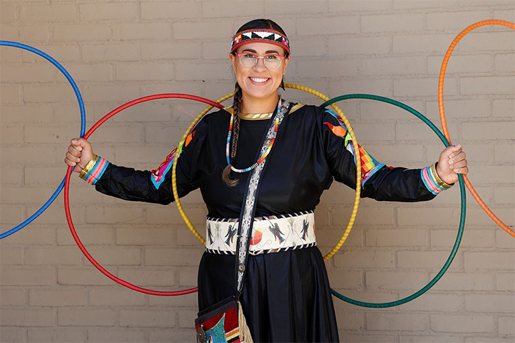 A smiling woman in traditional Indigenous regalia holds several colorful hoops in front of a wall.