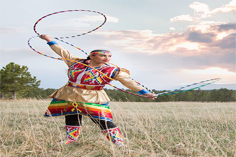 Indigenous woman in traditional regalia performing a hoop dance in a grassy field.