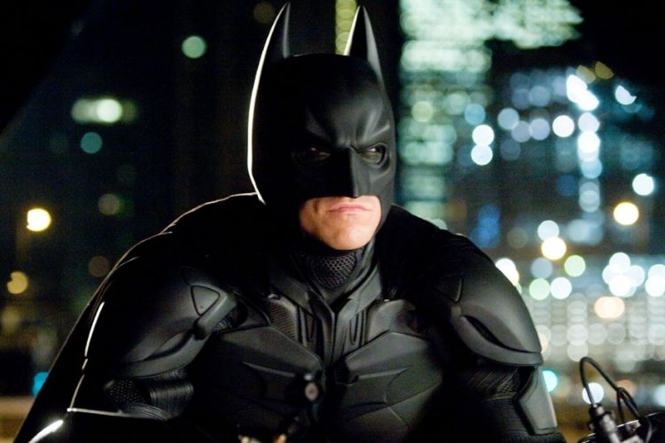 Photograph of Batman wearing his black tactical suit against a blurred city night background.