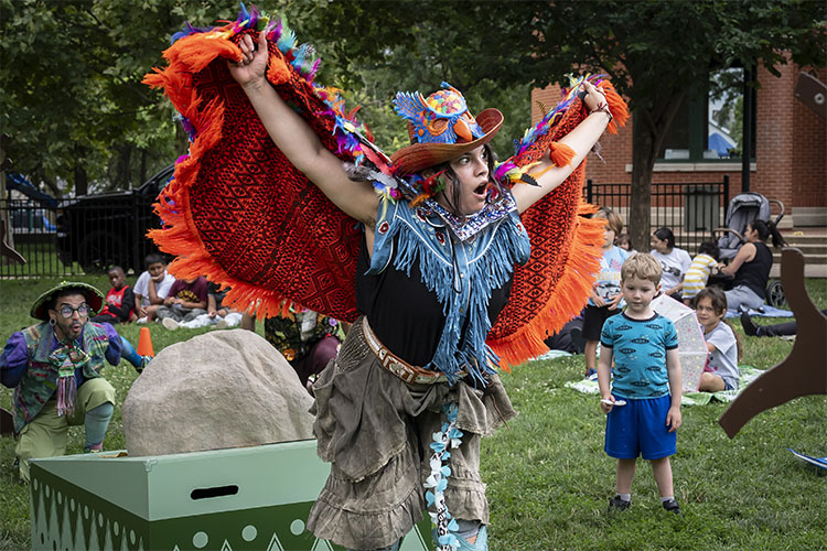 A costumed performer with large orange wings performs for children in a grassy park.