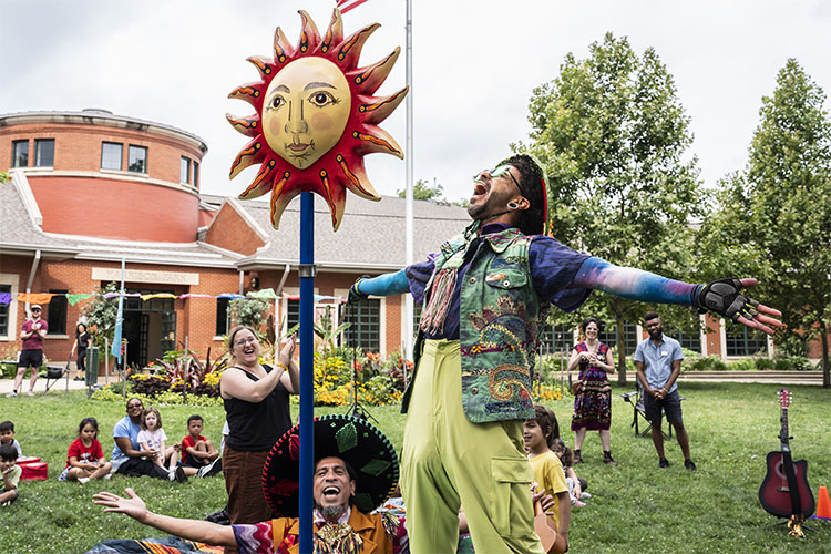 A performer with arms outstretched stands near a sun-shaped puppet before a park audience.