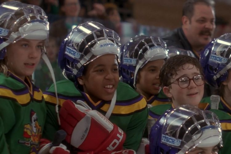 A group of young hockey players in green and purple jerseys watch a game from the bench.