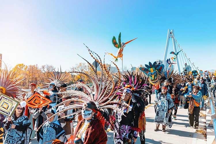 People in colorful traditional Aztec dance costumes with large feathered headdresses in a parade.