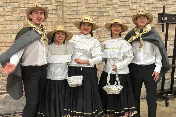 Five people in traditional folk costumes stand smiling before a brick wall.