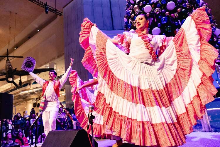 Performers in traditional attire dancing in front of a Christmas tree in a museum.