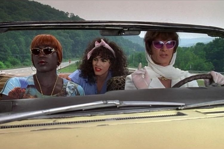 Three people in costume wearing sunglasses riding in a convertible on a mountain road.