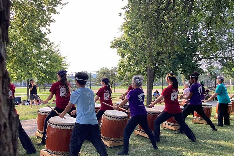 Group of people practicing Taiko drumming in a park, viewed from behind.