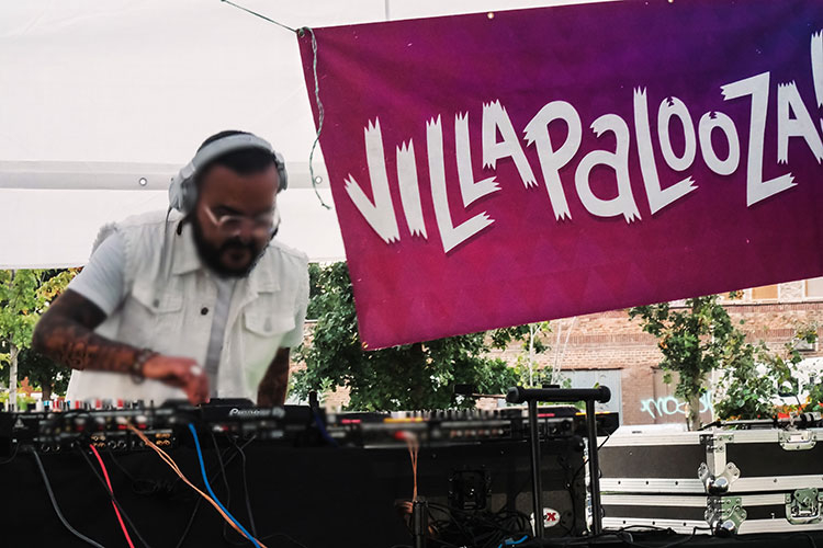 A DJ wearing headphones performs at an outdoor music festival with a "VILLAPALOOZA" banner.