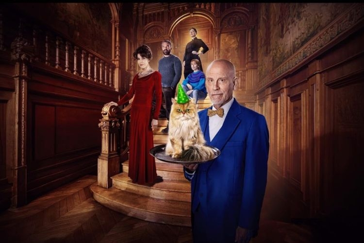 A man in a blue suit holds a tray with a fluffy cat wearing a party hat, before a group on stairs.