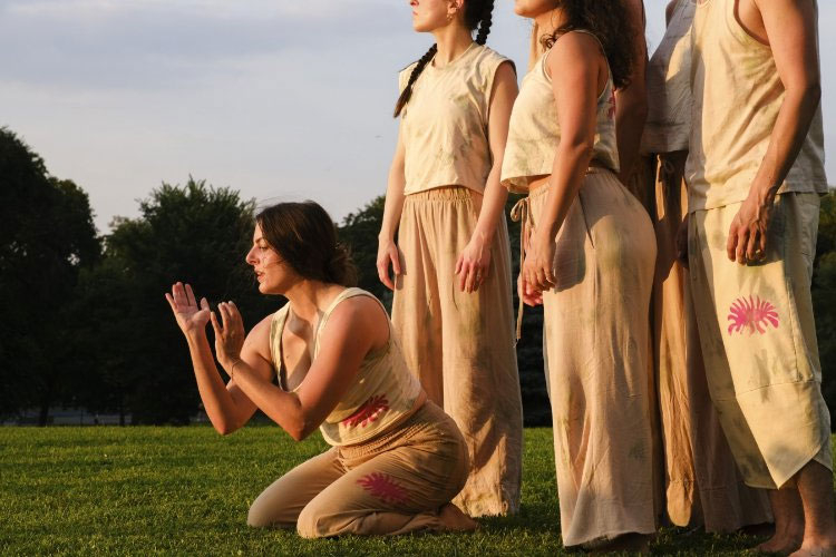 Photograph of dancers in matching beige outfits performing on a grassy field at sunset.