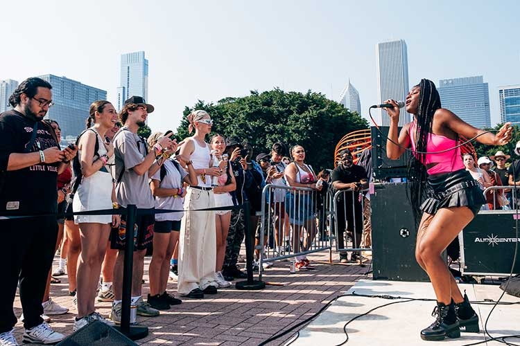 A female singer with long braids performs on a stage before an outdoor crowd with city buildings.