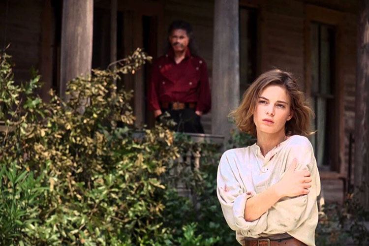 A young woman stands in the foreground, while a man watches from the porch of a rustic wooden house.